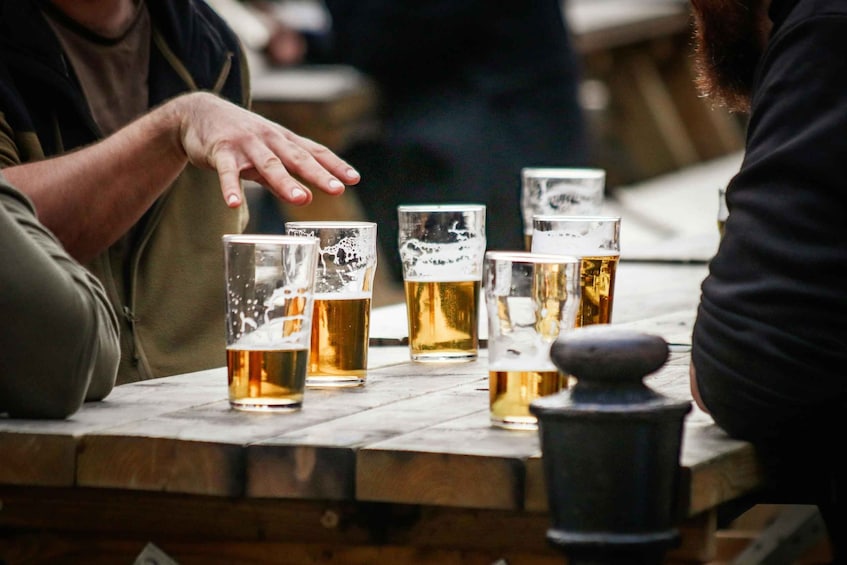A wooden table with several glasses of beer, a stone jug, and a person's hand reaching for a glass. 2