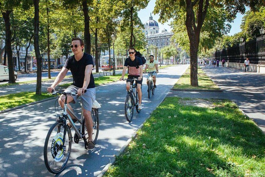 Tres personas andando en bicicleta por un sendero bordeado de árboles, con un edificio al fondo. 3