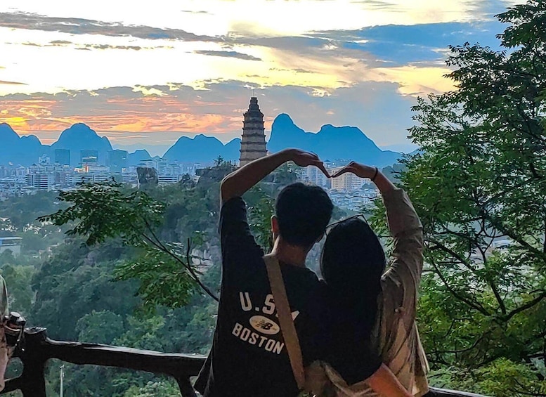 Zwei Personen formen mit ihren Händen ein Herz und blicken auf eine Stadtlandschaft mit Bergen und einem Turm. 1