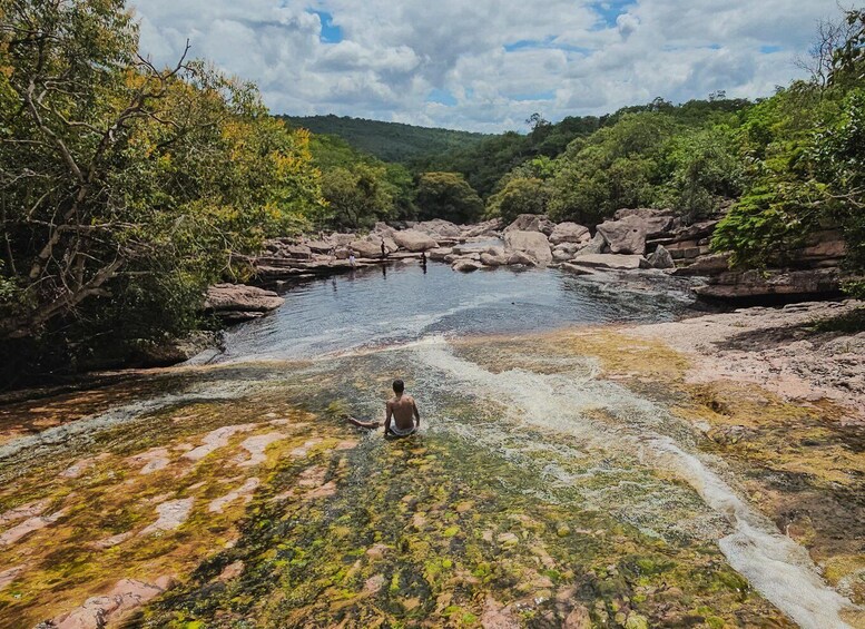 A person sitting by a clear, rocky river with lush greenery and hills in the background. 3