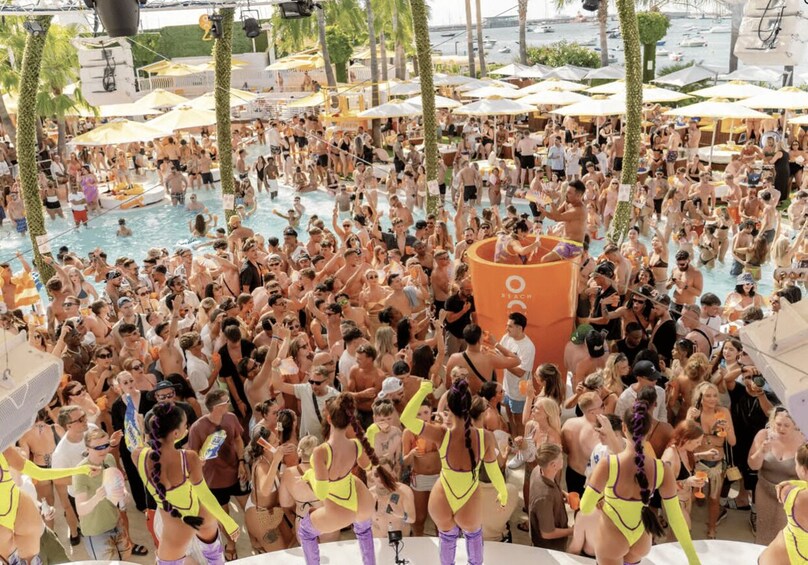 A crowded outdoor pool area with people swimming and socializing, surrounded by umbrellas and palm trees. 2