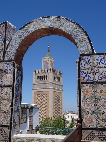 A view through a tiled archway towards a historic tower with intricate patterns and a green roof. 2