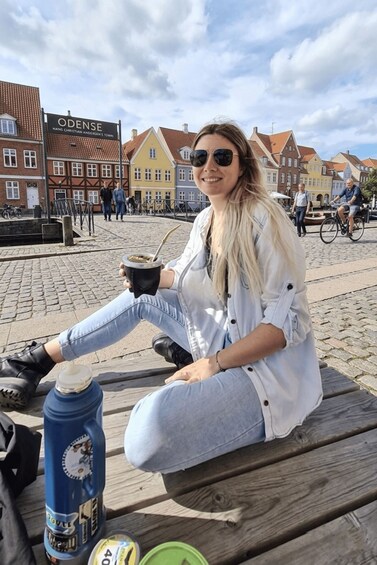 A person sitting on a wooden bench outdoors, holding a drink, with a blue water bottle and a green container nearby. 3