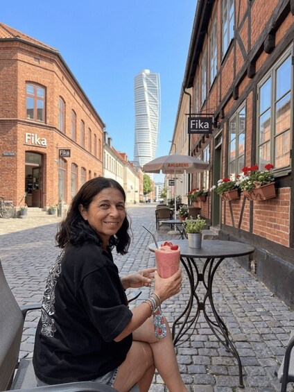 A person sitting on a bench holding a drink, with a brick building labeled 'Fika' and a modern skyscraper in the background. 2