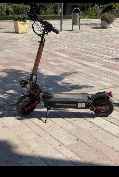 A stationary electric scooter on a paved surface with a planter and a trash bin in the background. 1