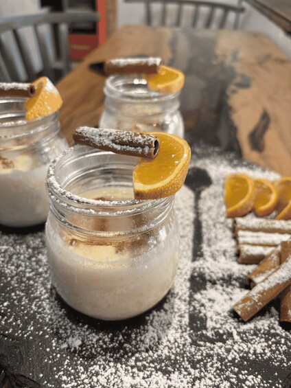 Three jars of a creamy dessert with cinnamon sticks and orange slices, dusted with powdered sugar on a dark surface. 2