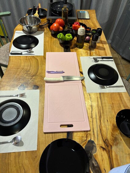 A wooden dining table set with black plates, a pink cutting board, and various kitchen utensils. 3
