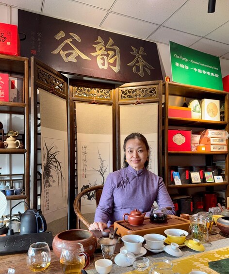 A person seated at a table with various tea utensils and containers, surrounded by shelves with tea products and a sign indicating a Chinese tea ceremony class. 1