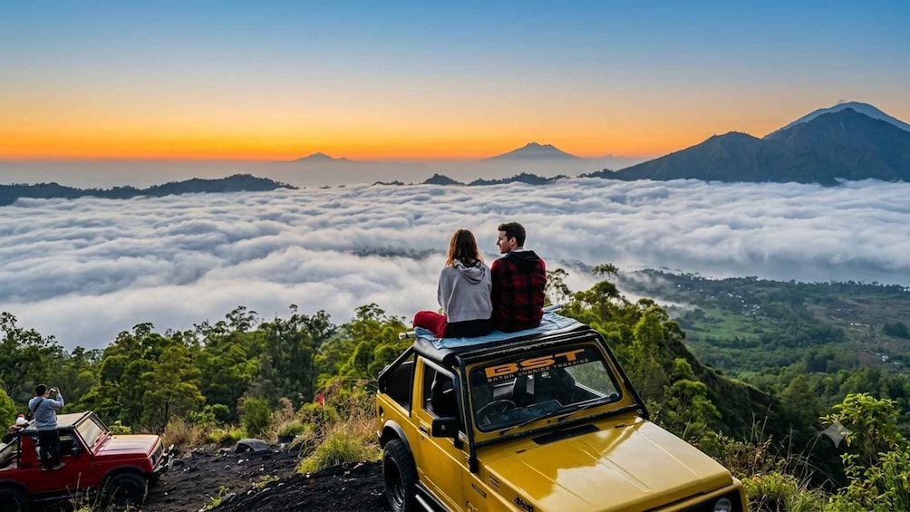 A yellow SUV with two people on top, overlooking a mountainous landscape with a sea of clouds below. 1