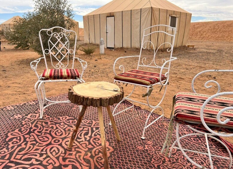 Two white metal chairs with patterned cushions, a round wooden table, and a patterned carpet in a desert setting with a tent in the background. 3