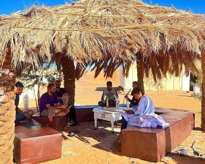 A group of people sitting under a thatched roof structure in a desert setting. 5