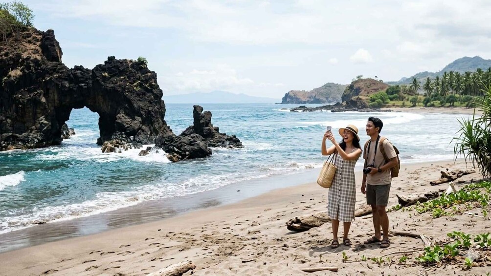 A couple taking a selfie on a beach with rocky cliffs and palm trees in the background. 1