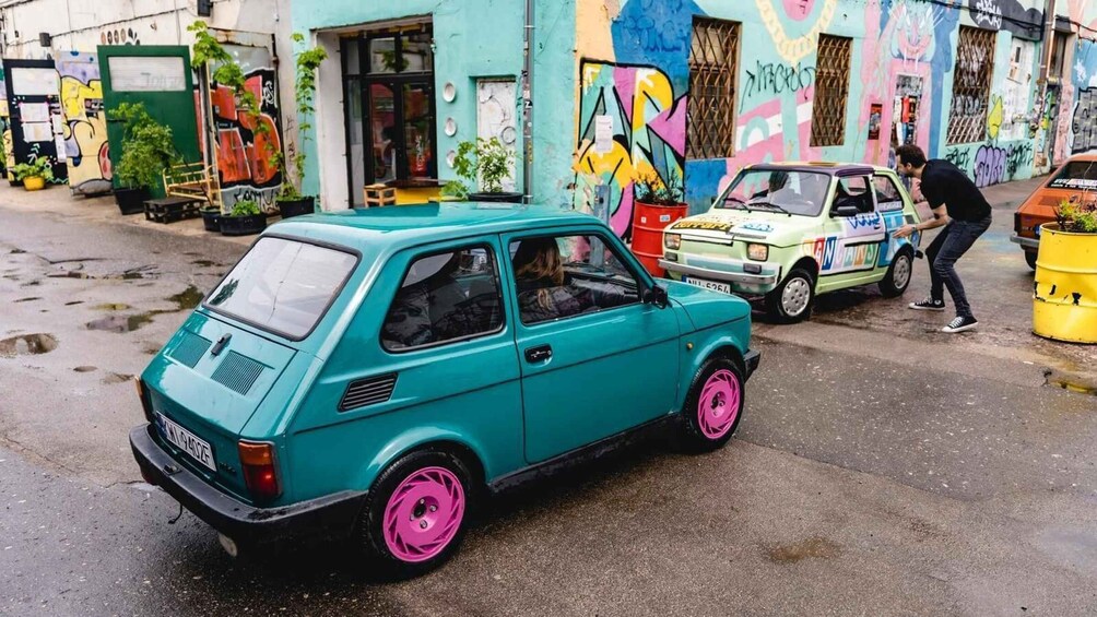 A teal car with pink rims parked on a street with colorful graffiti on the buildings. 3