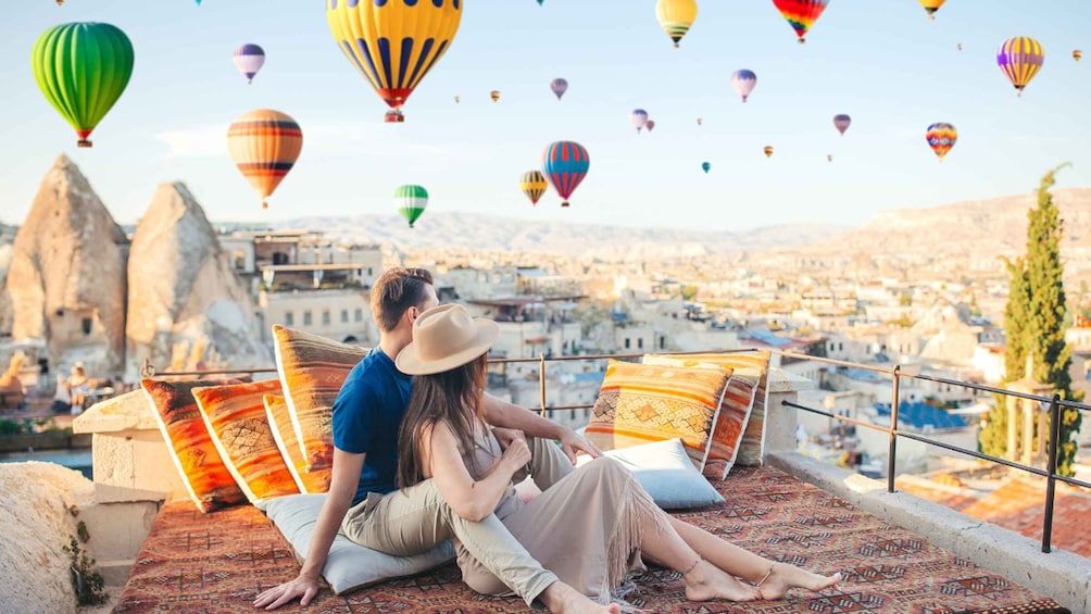 A couple sitting on a rooftop overlooking a cityscape with colorful hot air balloons in the sky. 1