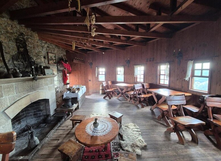 A rustic interior with a stone fireplace, wooden tables, and chairs, and a view of the outdoors through windows. 2