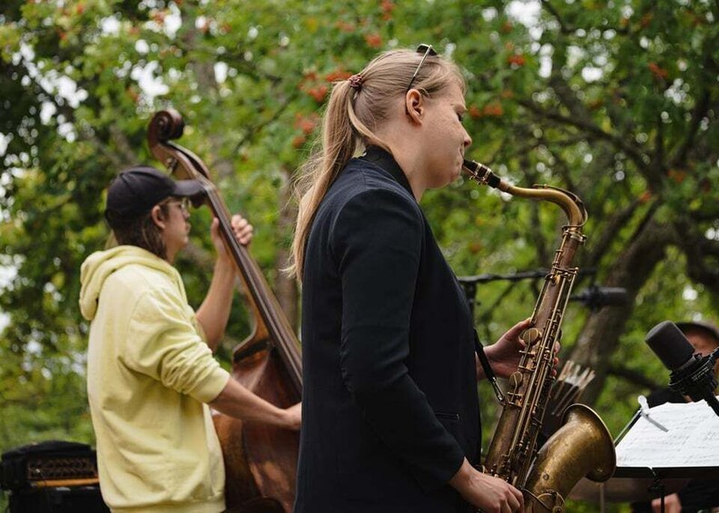 A person playing a brass instrument, likely a saxophone, outdoors with trees in the background. 5