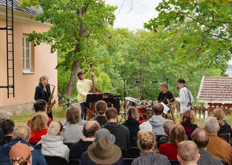A brass band performing outdoors with an audience seated in front. 1