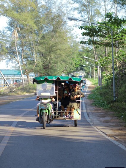 A green and white motorcycle cart with passengers on a road flanked by trees and streetlights. 5