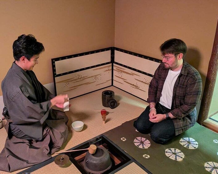 Two people sitting on tatami mats in a traditional Japanese room with a tea ceremony setup. 1