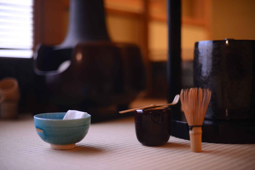 A traditional Japanese tea ceremony setup with a blue bowl, a dark ceramic cup, and a bamboo whisk on a tatami mat. 5