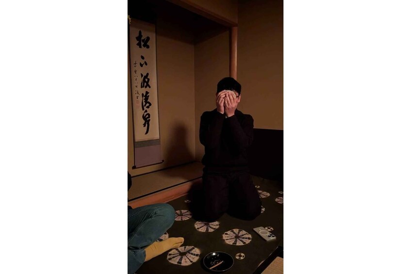 A person sitting on the floor with their hands covering their face, surrounded by tatami mats and a shoji screen with Japanese calligraphy. 4