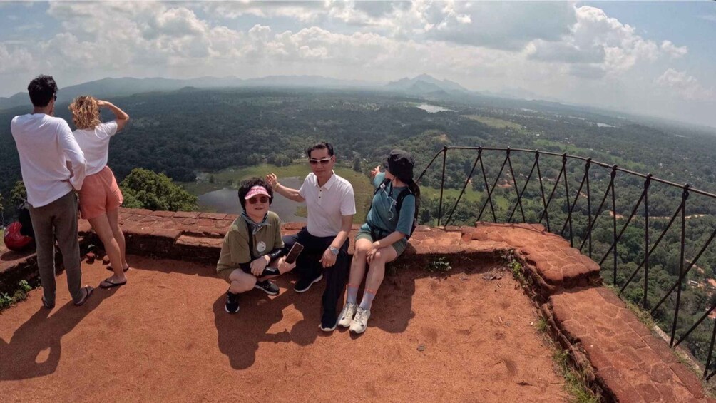 A group of people sitting on a red brick ledge overlooking a valley with mountains in the distance. 1