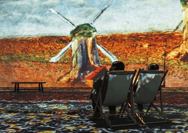 Two people seated on chairs facing a large painting of a windmill. 4