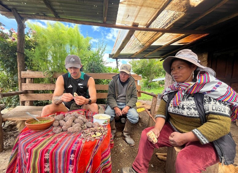 Three people sitting outdoors with a table of food and a colorful blanket. 2