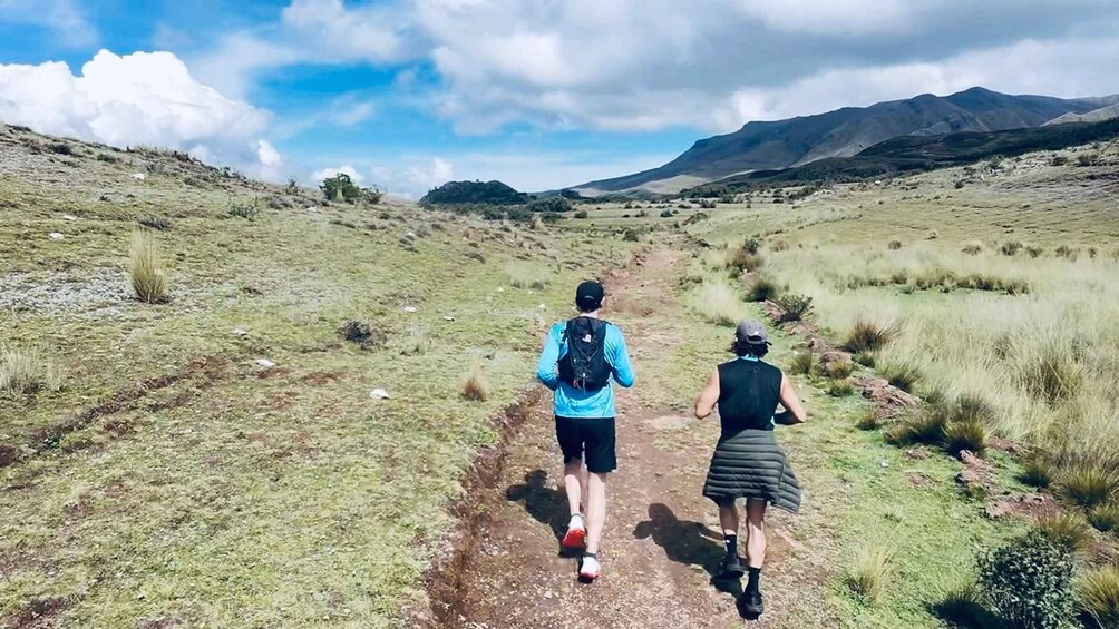 Two people walking on a dirt path in a grassy field with mountains in the background. 1