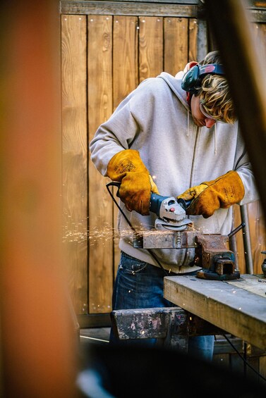 A person welding metal with sparks flying, wearing protective gloves and a hoodie, in front of a wooden background. 2