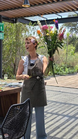 A person holding a bouquet of flowers, wearing an apron with a logo, standing under a wooden pergola with greenery in the background. 3
