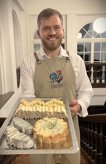 A person holding a tray of various cheeses in a room with arched windows and wooden furniture. 3