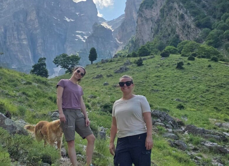 Two people standing on a grassy hillside with a dog, mountains in the background. 1