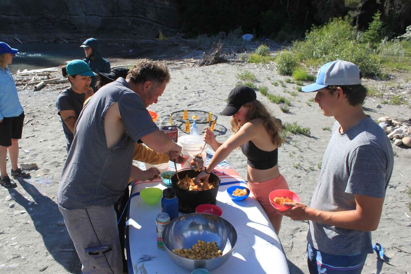 A group of people gathered around a table with food and drinks on a beach. 1