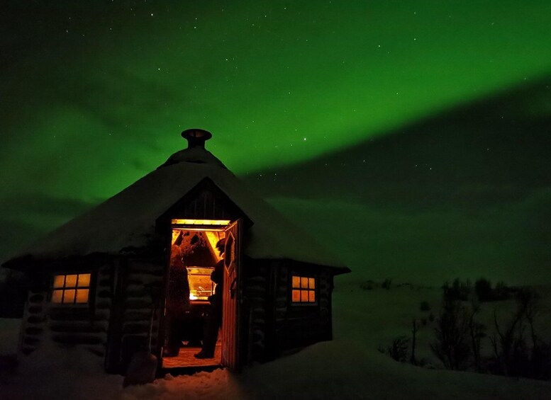 A log cabin with a lit interior, surrounded by snow, under a green aurora. 3