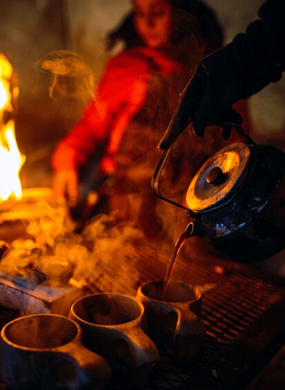 A person pouring tea into a cup from a teapot over a fire. 1