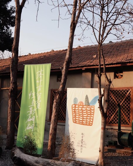 A building with a tiled roof and two banners, one green and one white, with text and a basket illustration. 2
