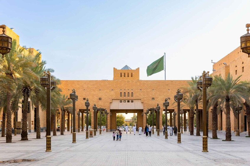 A large, historic building with a prominent entrance, palm trees lining the walkway, and a flag flying atop. 1