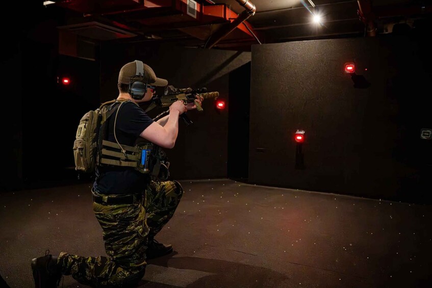 A person in tactical gear aiming a rifle in a dimly lit indoor shooting range. 3