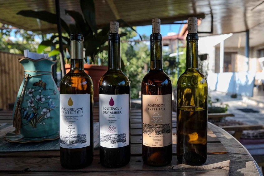 Four bottles of wine on a wooden table under a covered area with plants in the background. 1
