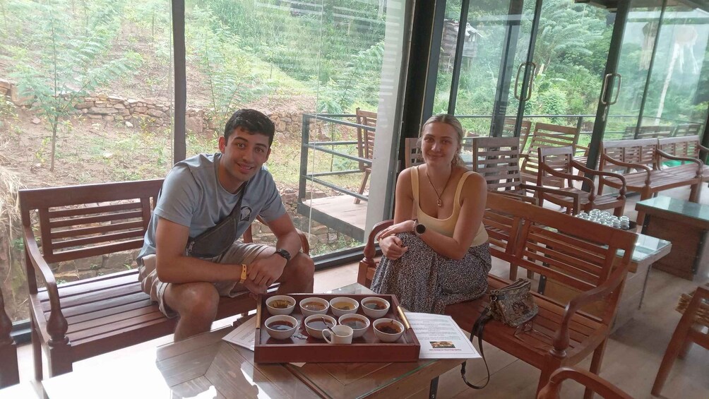 Two people seated at a table with a tray of cups, in a room with large windows overlooking a garden. 4