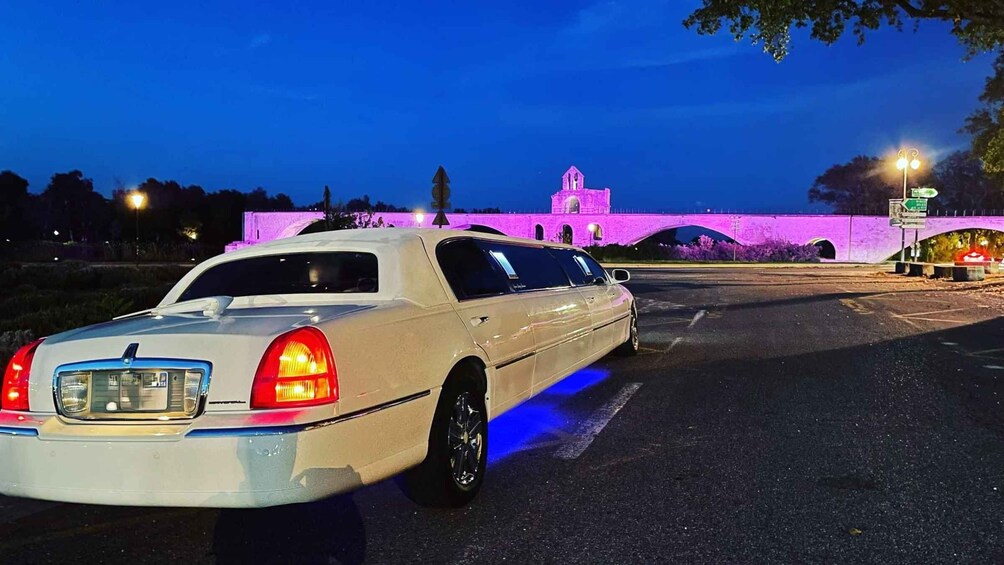 A white luxury sedan parked in a lot with a bridge and illuminated building in the background. 1