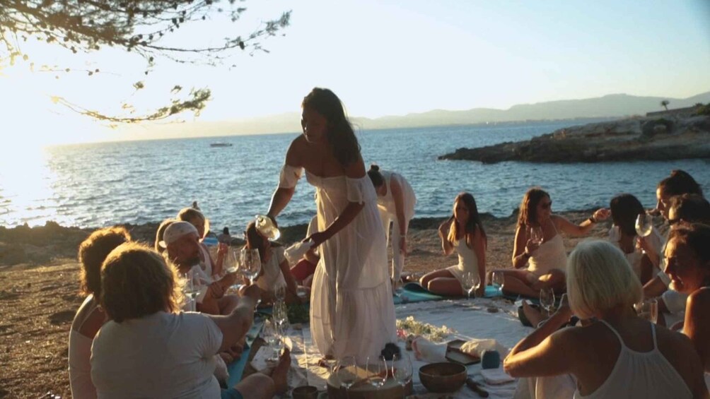 A group of people sitting on the beach at sunset, engaged in a picnic with wine glasses and a table set with food. 2