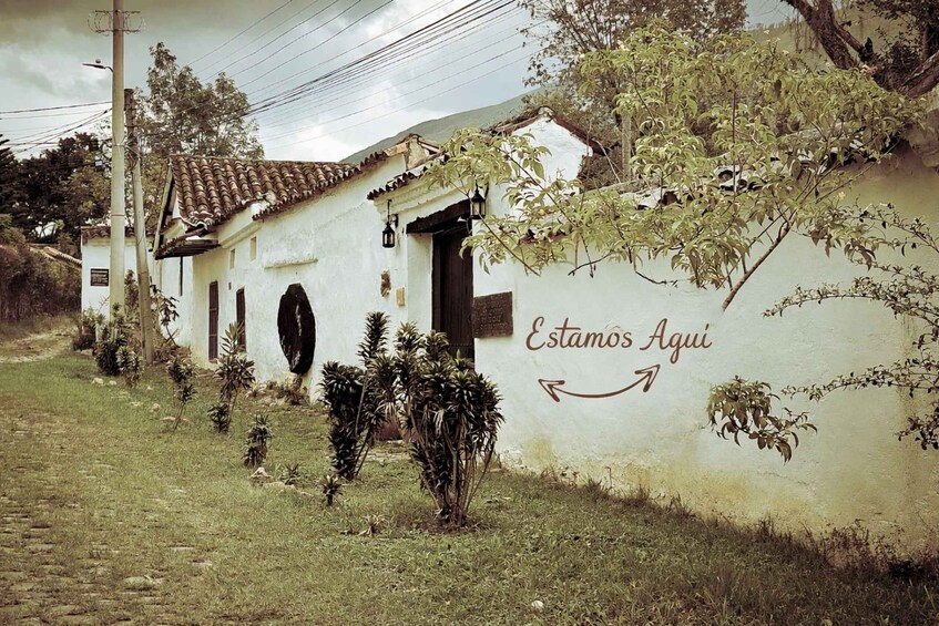 A white building with a tiled roof, a sign reading 'Estamos Aqui,' and a small garden with plants. 1