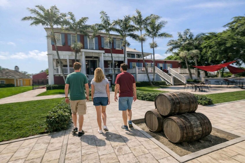 Three people walking towards a building with a red and white facade, palm trees, and wooden barrels in the foreground. 2