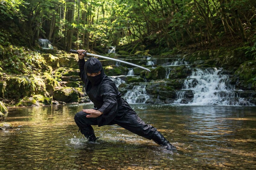 A person in black attire performing a water-based martial arts move in a forested area with a waterfall. 2