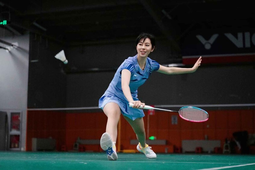 A person playing badminton in an indoor court with a net, red walls, and a visible logo. 3
