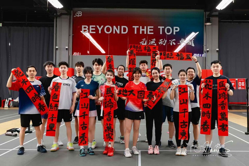 A group of people holding red banners with Chinese characters, standing in an indoor sports facility with a large red banner in the background that reads 'BEYOND THE TOP.' 2