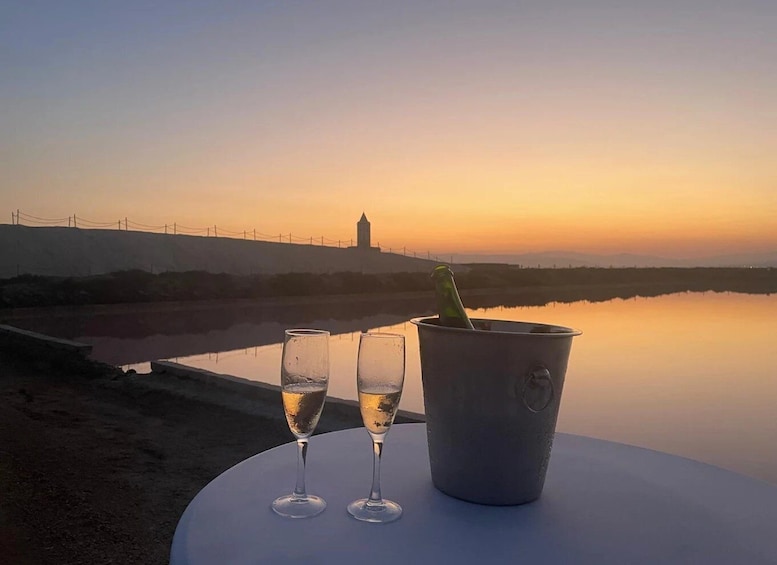 A sunset over a calm body of water with a distant tower silhouette, a table with two glasses and a champagne bottle. 2