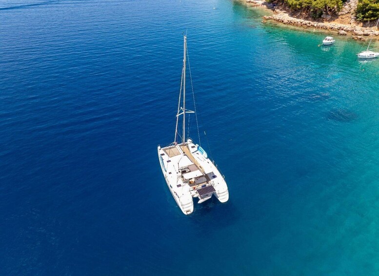 A white sailboat anchored in a clear blue sea with a rocky shoreline and a small boat in the background. 1